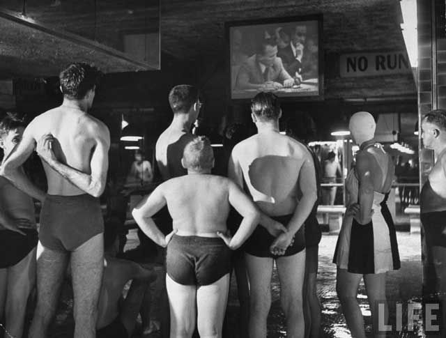 "Fascinated group of swimmers at indoor swimming pool, watching large TV screen showing Russian Amb. to the UN Jacob Malik filibustering to tie the Security Council in parliamentary knots." August 1950